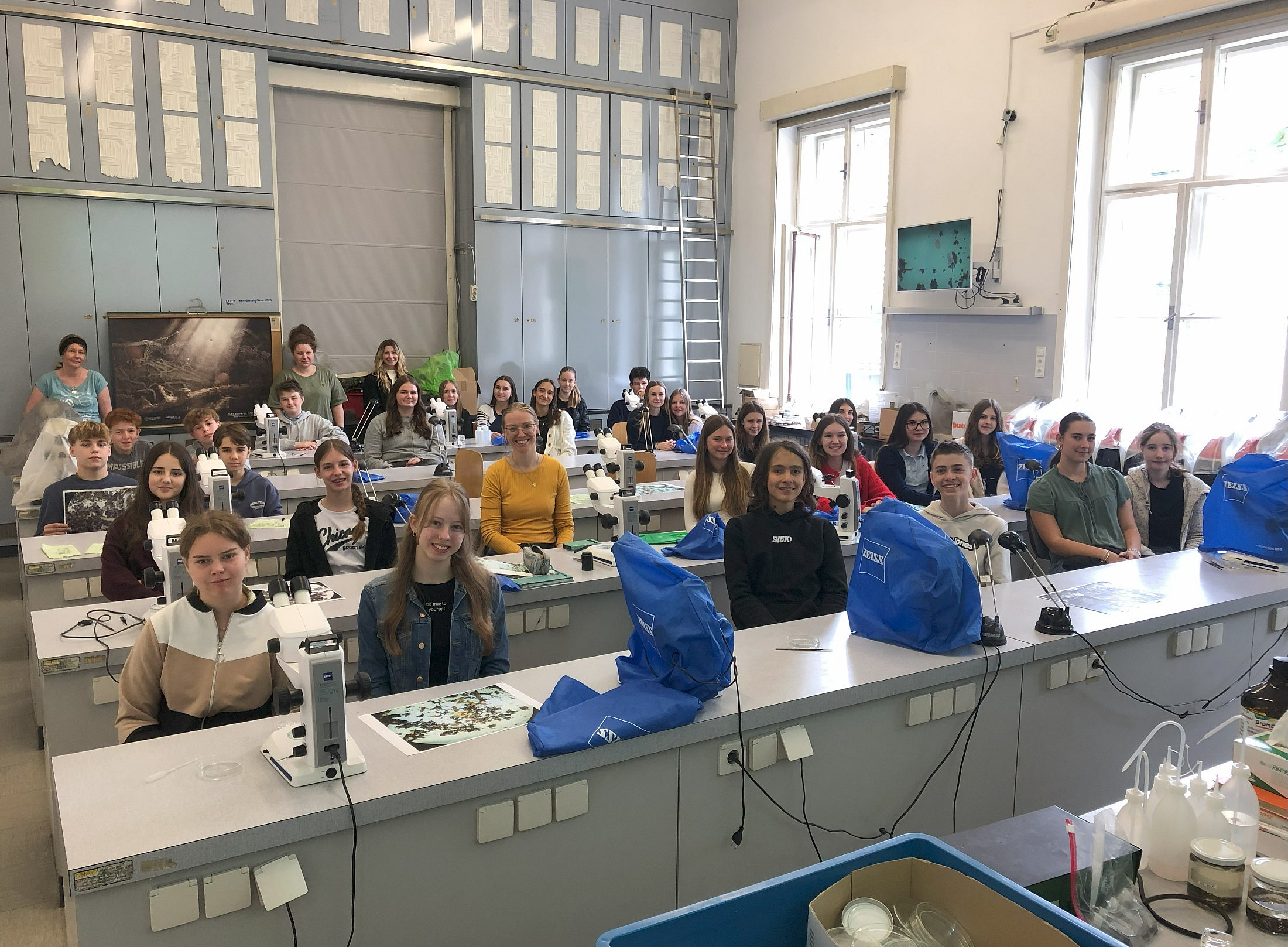 Eine Gruppe von Schülern in einem deutschen Schullabor sitzt an ihren Schreibtischen und lächelt für ein Foto, auf denen blaue Taschen mit naturwissenschaftlichen Utensilien auf jedem Schreibtisch stehen. Vor ihnen steht ein elektrisches Mikroskop, das Licht in eine kleine Plastikbox mit organischem Material strahlt. 