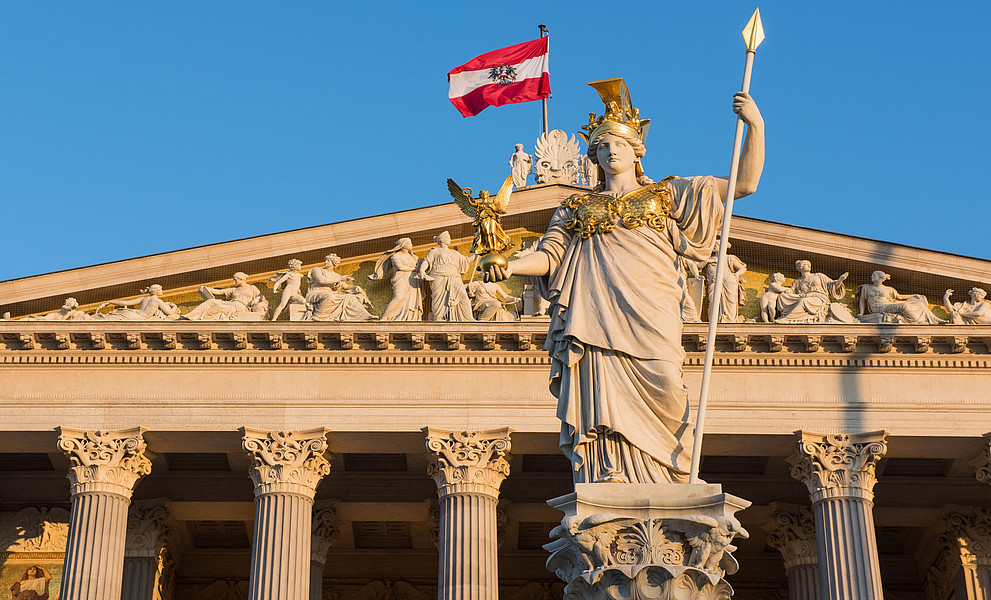 Ein Foto der österreichischen Nationalflagge, die über einer eleganten Statue der Justitia vor dem österreichischen Parlament weht, einem prächtigen Gebäude in Wien, das mit seinen Säulen und Bögen an die antike griechische Architektur erinnert und mit goldenen Statuen geschmückt ist. 