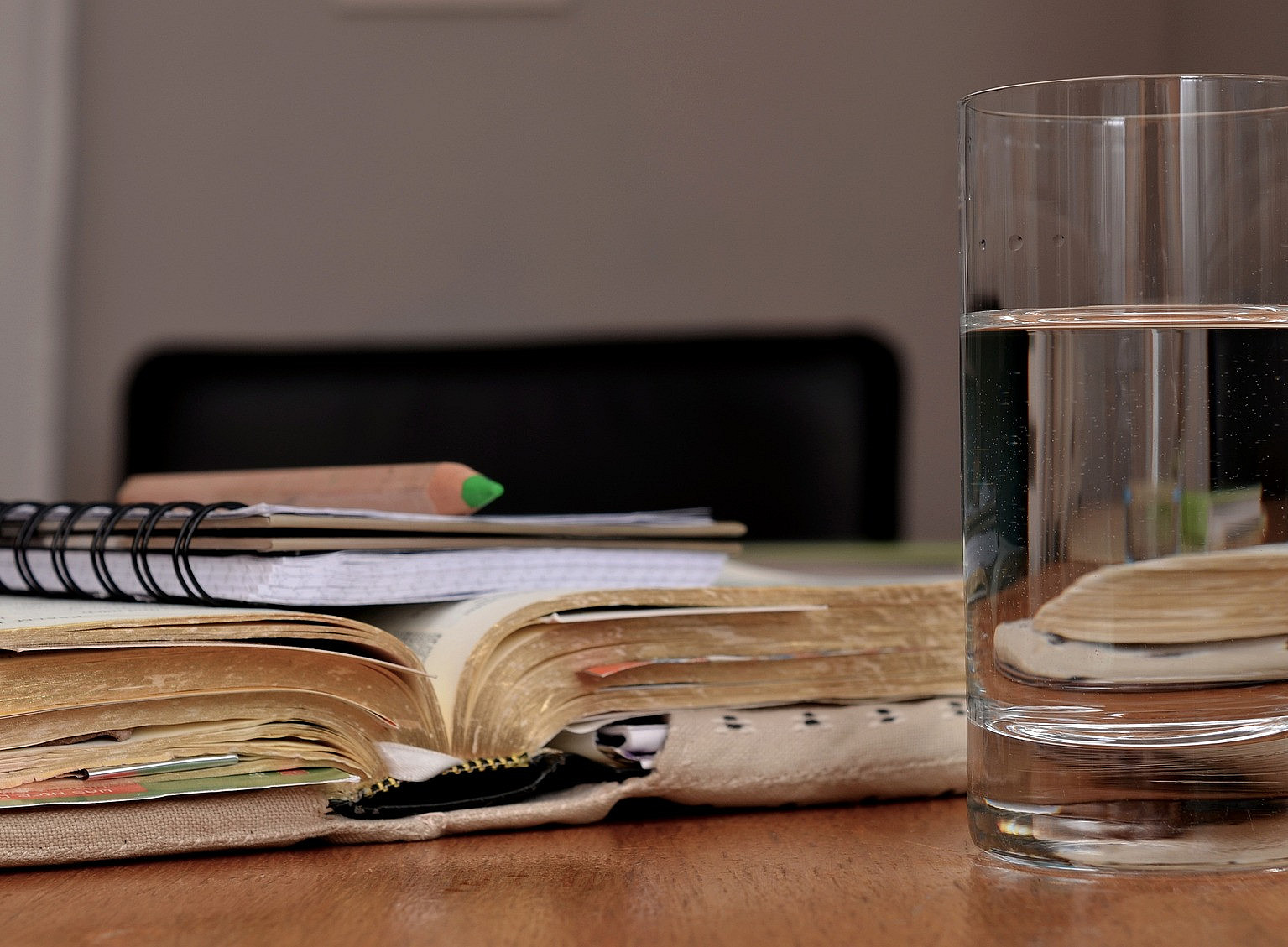 Ein Buch mit Stoffumschlag liegt aufgeschlagen auf einem Tisch. Auf dem Buch liegt ein Notizblock mit einem grünen Buntstift. Im Vordergrund steht ein Glas Wasser. ©Pixabay (congerdesign)