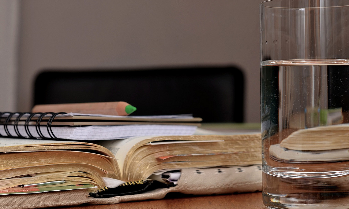 Ein Buch mit Stoffumschlag liegt aufgeschlagen auf einem Tisch. Auf dem Buch liegt ein Notizblock mit einem grünen Buntstift. Im Vordergrund steht ein Glas Wasser.