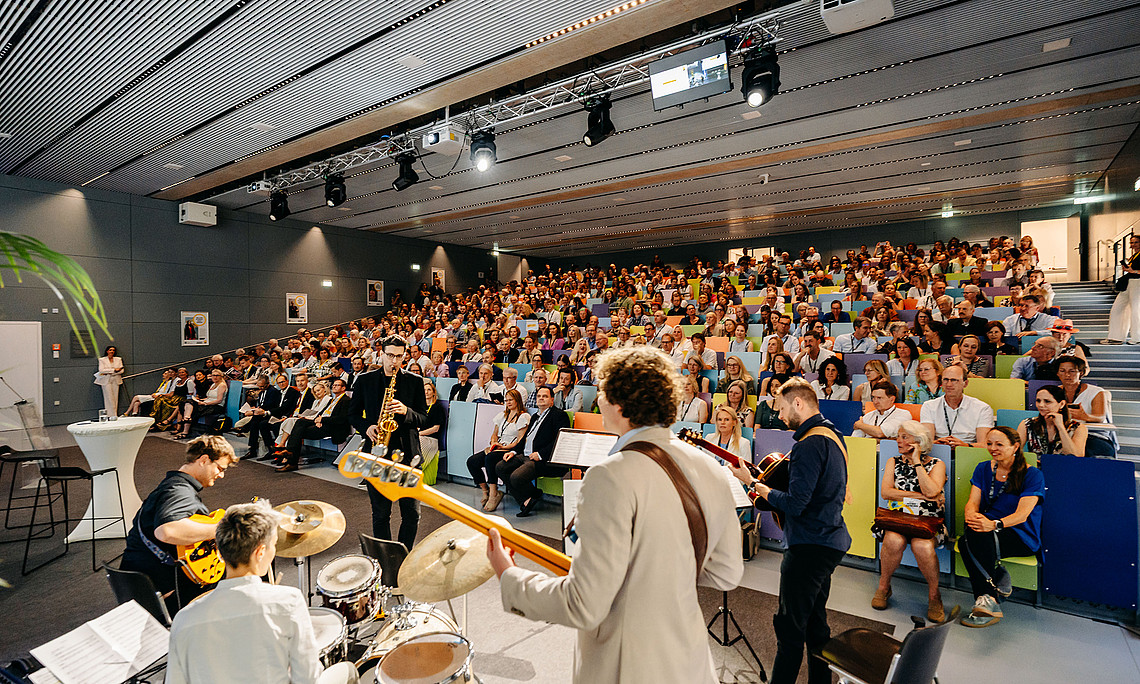 Ein großer Hörsaal der voll mit Personen ist. Im Vordergrund sitzt man eine Band