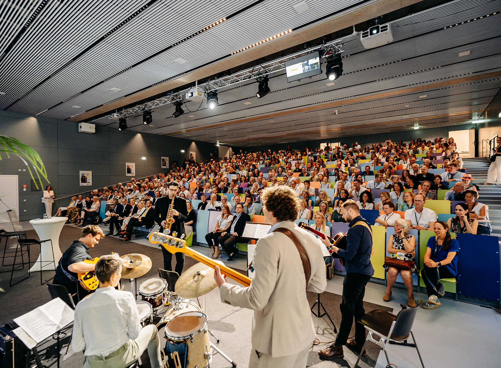 Ein großer Hörsaal der voll mit Personen ist. Im Vordergrund sitzt man eine Band ©© Nadja Fuchs 