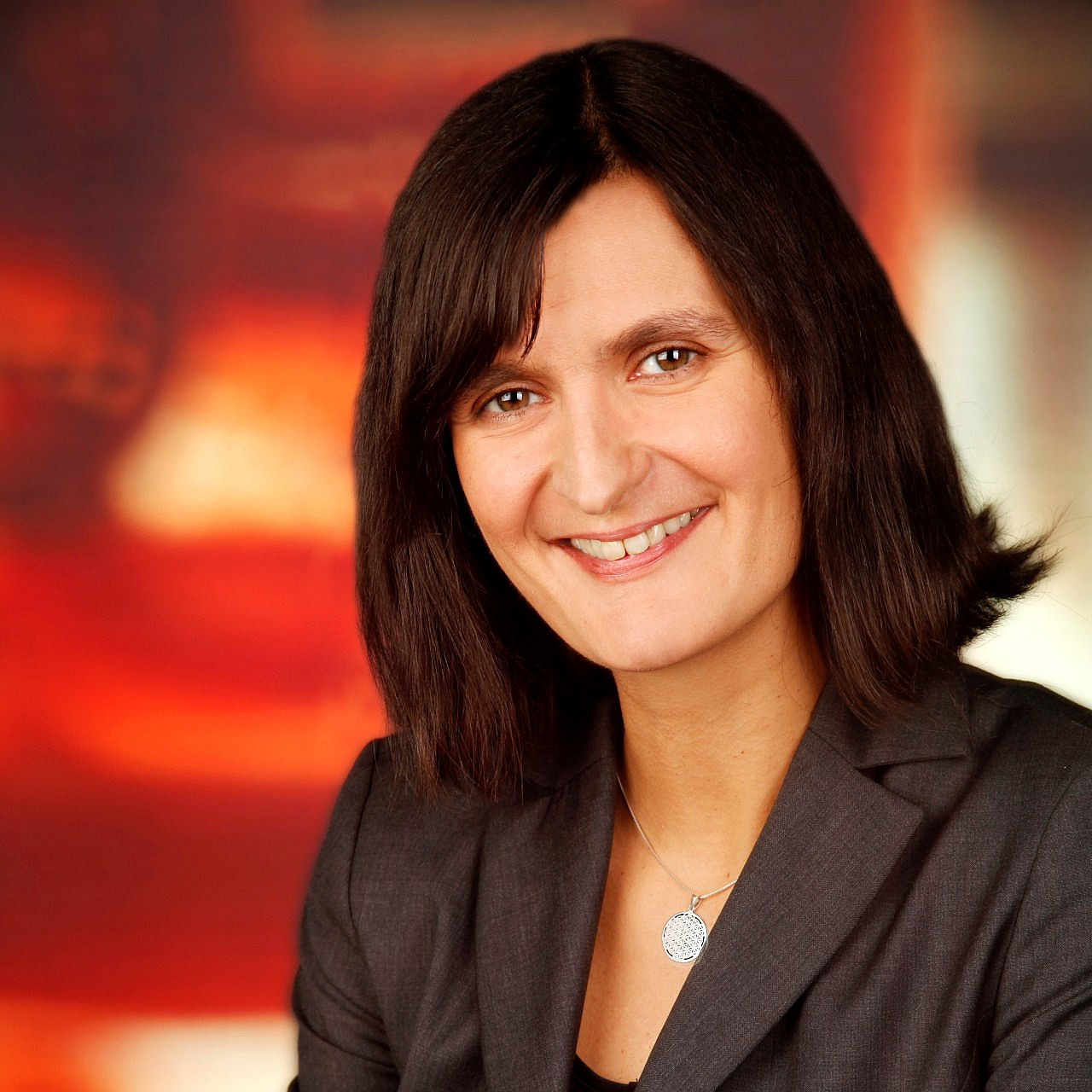 eine Frau mit braunen schulterlangen Haaren in einem grauen Blazer, sie hat die Arme verschränkt und lächelt in die Kamera