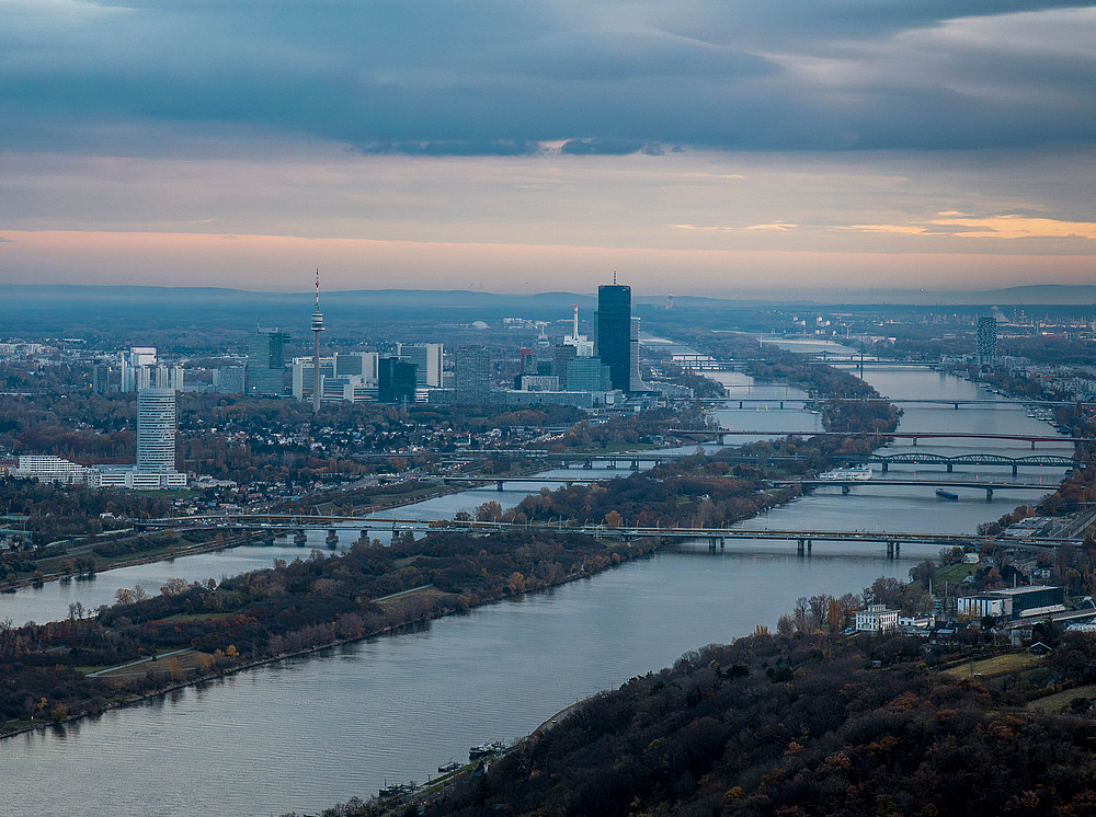 A panoramic view of the city of Vienna along the Danube at sunset, with modern skyscrapers, bridges and tree-lined banks. The sky shows a colour gradient from blue to orange. ©Manuel Madaini