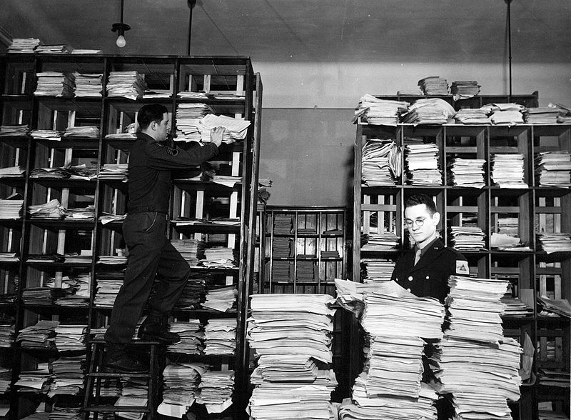 Schwarz-weiß-Fotografie eines Archivs mit hohen Regalen voller Aktenstapel. Ein Mann steht auf einer Leiter und sortiert Dokumente in ein Regal, während ein anderer Mann in Uniform neben einem großen Stapel Akten steht.“ 