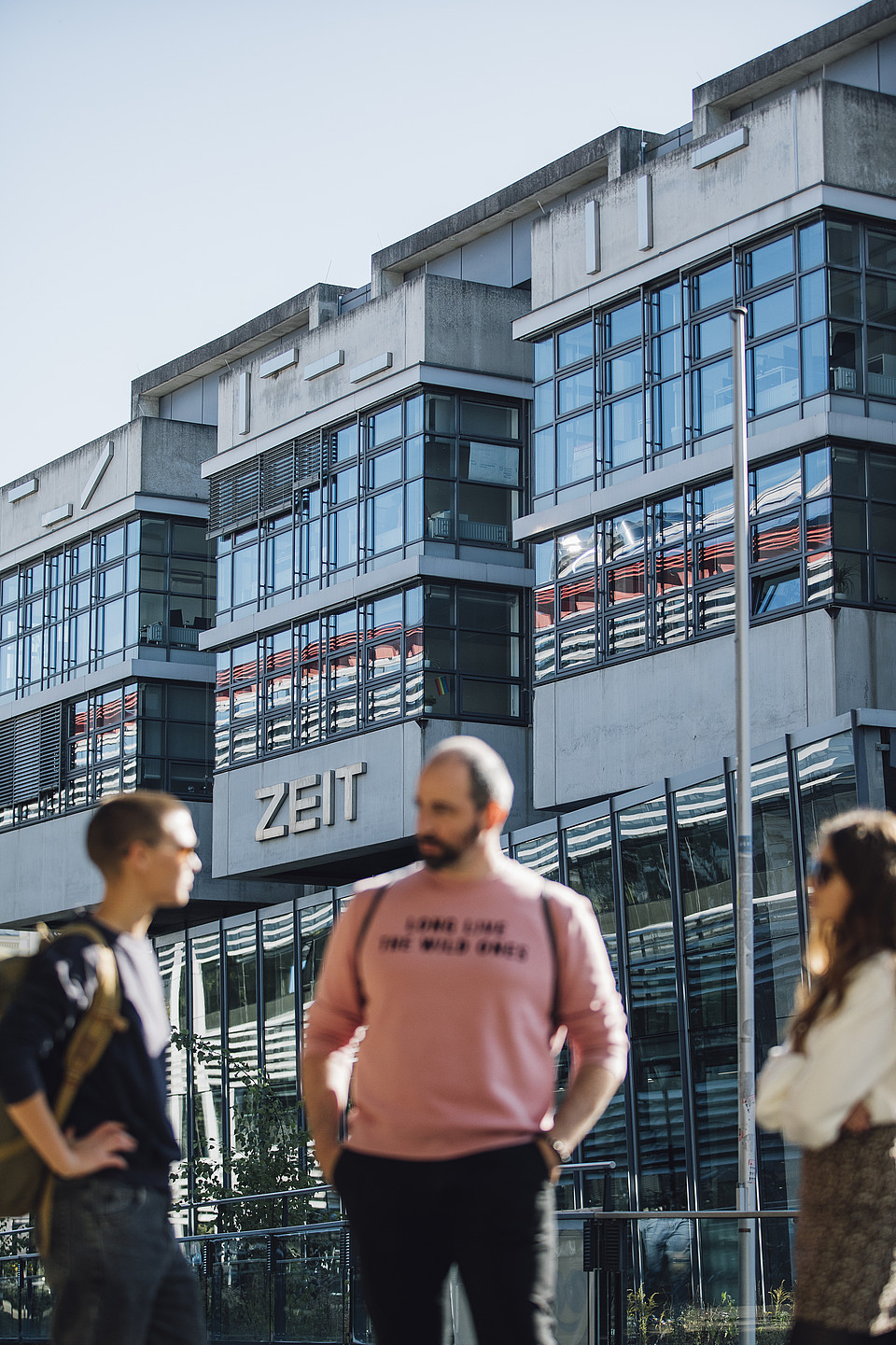 Studierende unterhalten sich am Campus, dahinter ist scharf ein Gebäude mit vielen Fenstern zu sehen, der Schriftzug "ZEIT" steht auf dem Gebäude