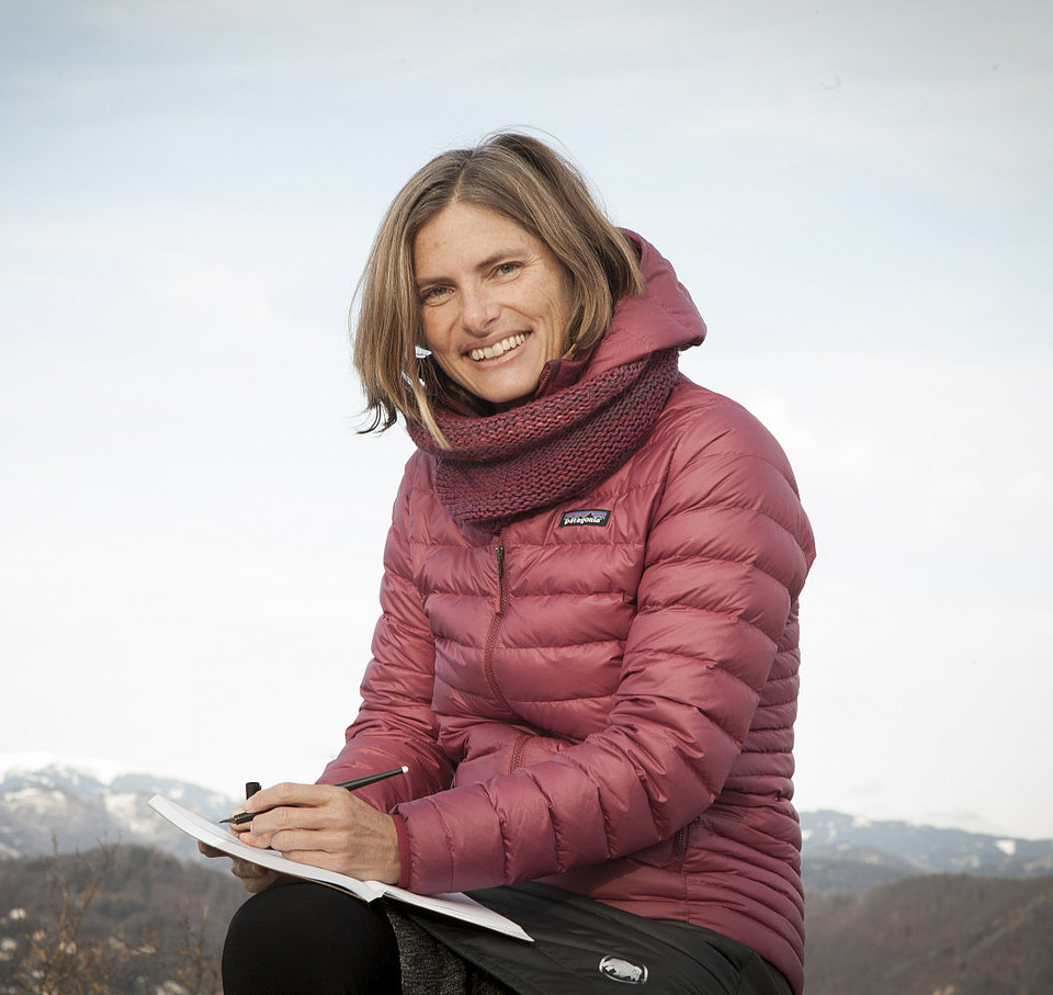 Fotografie von Michaela Nutz, USW Geo-Absolventin, im Hintergrund Berge, in der Hand hält sie einen Block mit Stift und lacht in die Kamera.