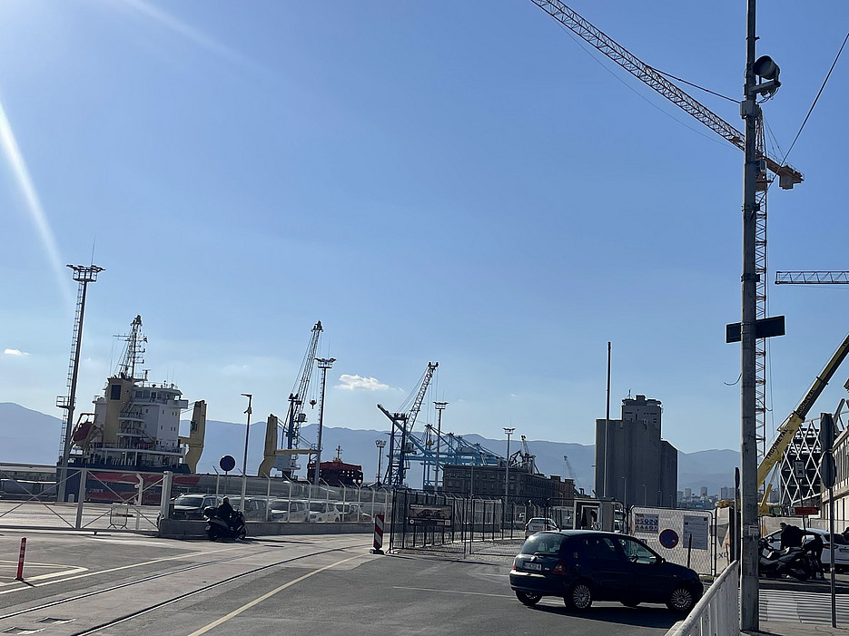 A busy port with large cranes, a docked ship, and industrial buildings against a clear blue sky. Cars are parked nearby, suggesting an active area. ©Milka Car