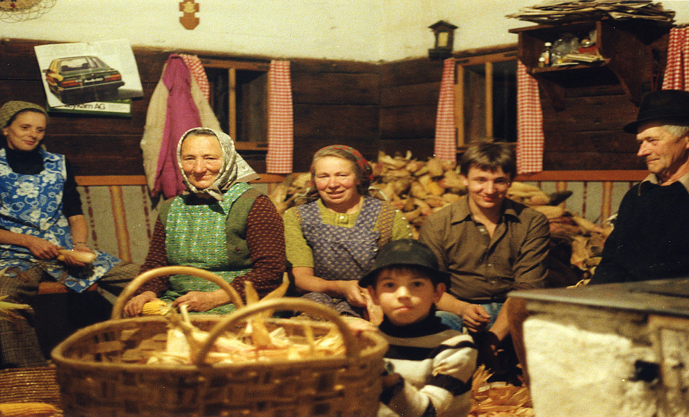 Bäuerliche Familie, sechs Personen aus drei Generationen, in einer Stube mit einem Korb voller ungeschälter Maiskolben.