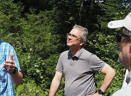 Foto von drei Männern im Wald. Einer trägt eine Brille, hat kurzes graues Haar und trägt ein graues Polohemd, während er mit dem anderen spricht. Der zweite Mann ist von hinten zu sehen und hat eine weiße Mütze auf dem Kopf. Vor ihnen ist hohe grüne Vegetation zu sehen, der Himmel ist klar. ©Nationalpark Gesäuse/Wölger