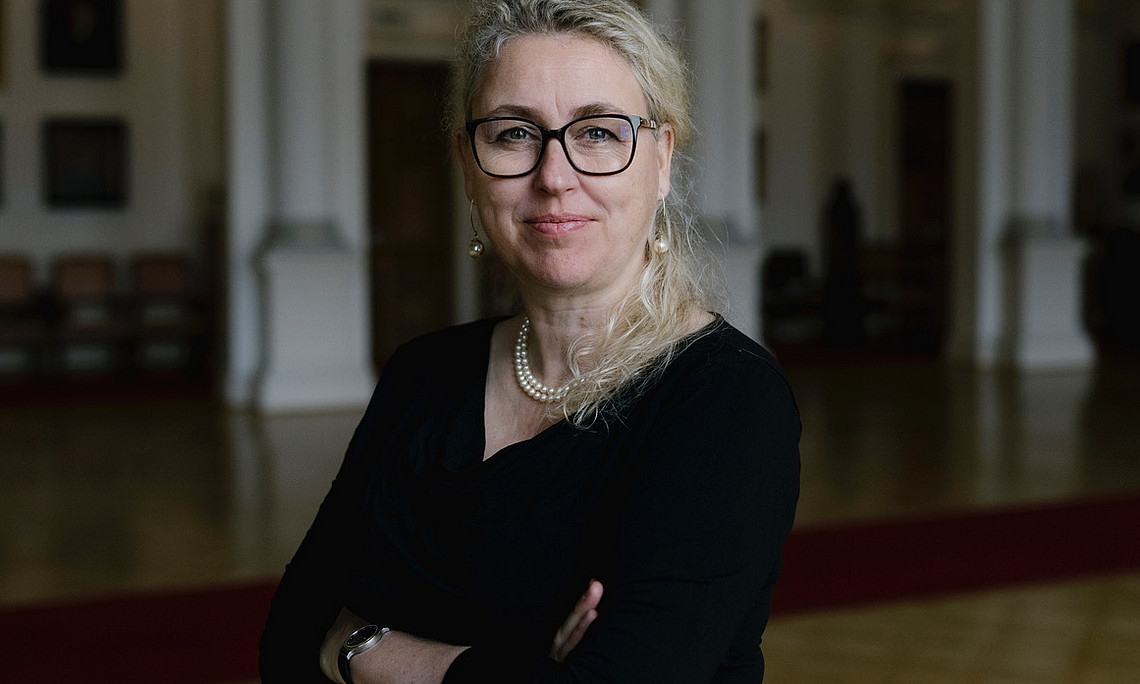 Porträt einer lächelnden Frau mit Brille und Perlenkette, blondem Pferdeschwanz und schwarzem Hemd, die in der Aula der Universität Graz steht. Der Hintergrund ist verschwommen, was eine stimmungsvolle Atmosphäre schafft. 