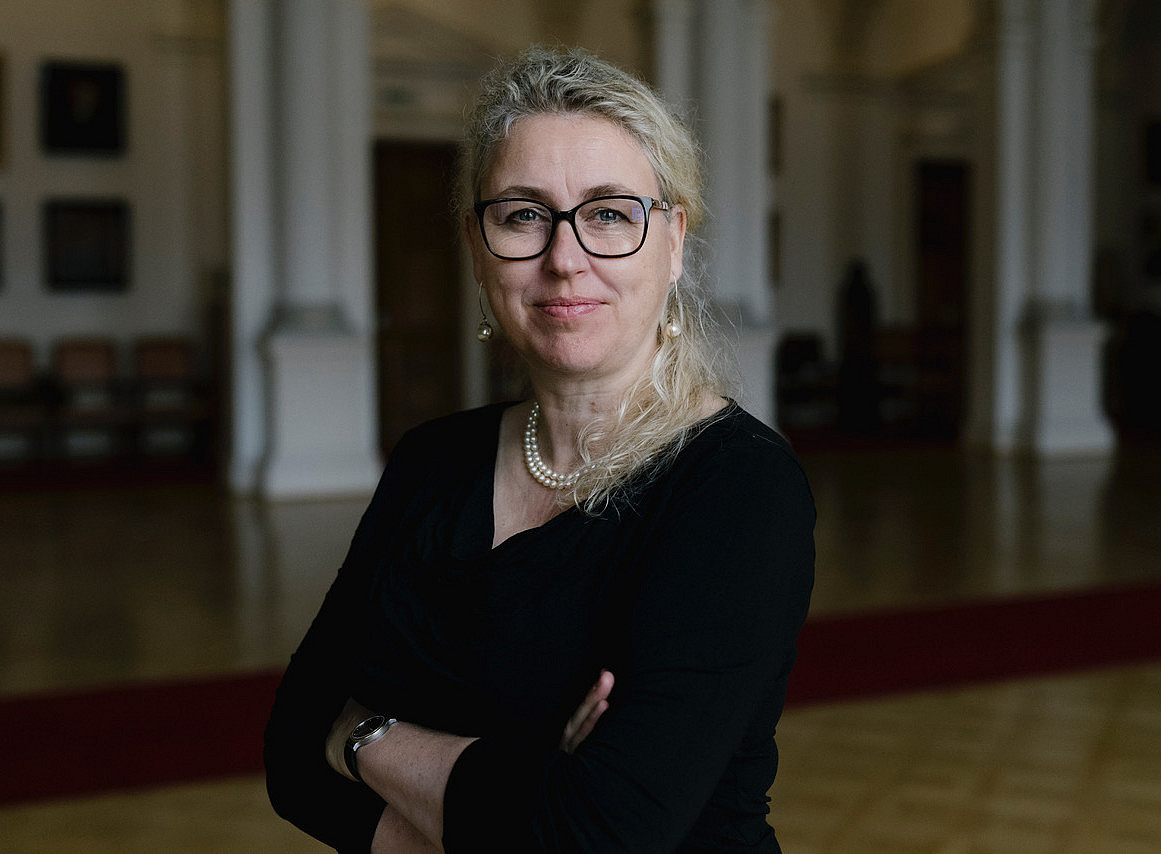 Porträt einer lächelnden Frau mit Brille und Perlenkette, blondem Pferdeschwanz und schwarzem Hemd, die in der Aula der Universität Graz steht. Der Hintergrund ist verschwommen, was eine stimmungsvolle Atmosphäre schafft. 