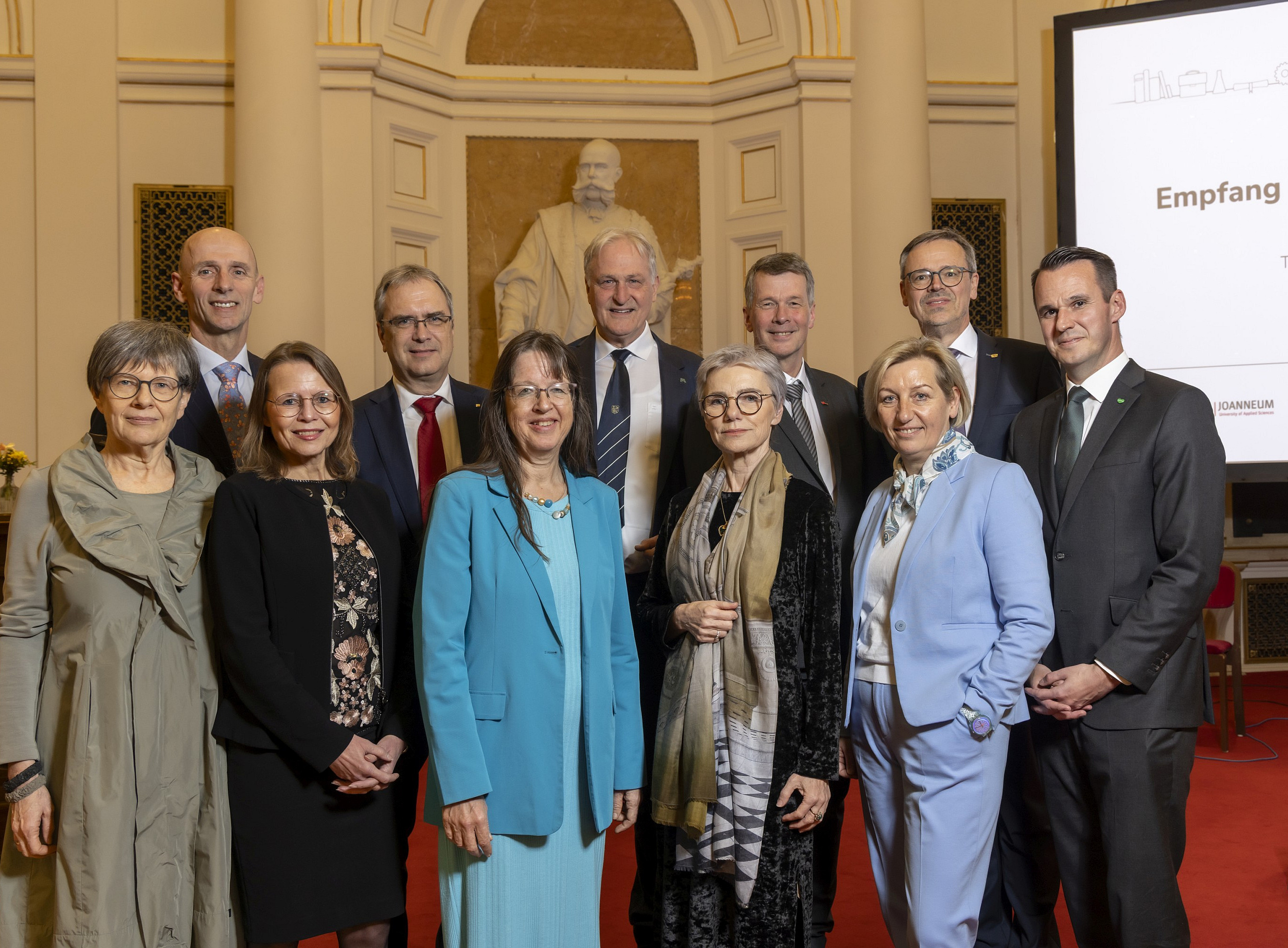 Eine Gruppe elegant gekleideter Personen steht dicht beisammen in einem festlich wirkenden Saal mit hohen, cremefarbenen Wänden und Säulen. Im Hintergrund ist eine große weiße Statue vor einer goldenen Nische zu sehen. Rechts steht ein hoher Bildschirm mit weißer Präsentationsfolie und mehreren Logos im unteren Bereich. Der Boden ist mit rotem Teppich ausgelegt, und die Personen tragen überwiegend Anzüge, Kostüme und festliche Kleider. ©© Helmut Lunghammer