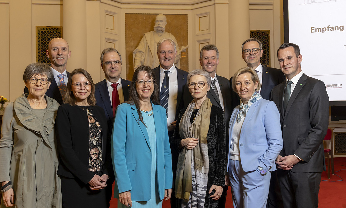 Eine Gruppe elegant gekleideter Personen steht dicht beisammen in einem festlich wirkenden Saal mit hohen, cremefarbenen Wänden und Säulen. Im Hintergrund ist eine große weiße Statue vor einer goldenen Nische zu sehen. Rechts steht ein hoher Bildschirm mit weißer Präsentationsfolie und mehreren Logos im unteren Bereich. Der Boden ist mit rotem Teppich ausgelegt, und die Personen tragen überwiegend Anzüge, Kostüme und festliche Kleider.