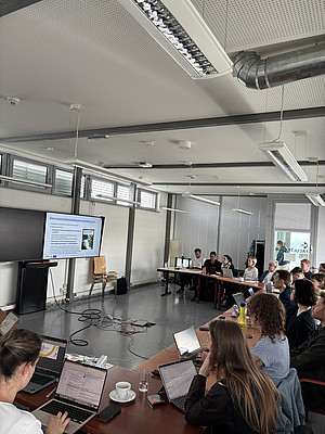 Rund 15 Erwachsene sitzen mit Laptops an Tischen und verfolgen eine Präsentation auf einem Bildschirm mit Folien zur österreichischen Klimawandelanpassungsstrategie. ©Paul Schweighofer