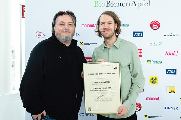 Bienenforscher Thomas Schmickl und Ex-Formel-1-Fahrer Sebastian Vettel mit der Ehren-Urkunde für den Bienenforschungspark vor einer Logo-Wand ©Uni Graz/Konstantinov