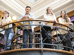 Vier Studierende stehen auf der Galerie des historischen Lesesaals mit Blick hinab in die Kamera. ©Uni Graz/Tzivanopoulos