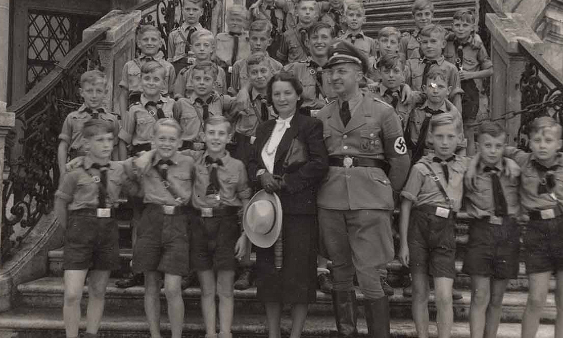 Gruppe von Jungen in Uniform, eine Frau und ein Mann in NS-Uniform stehen auf einer Treppe vor einem Gebäude, alle blicken in die Kamera.