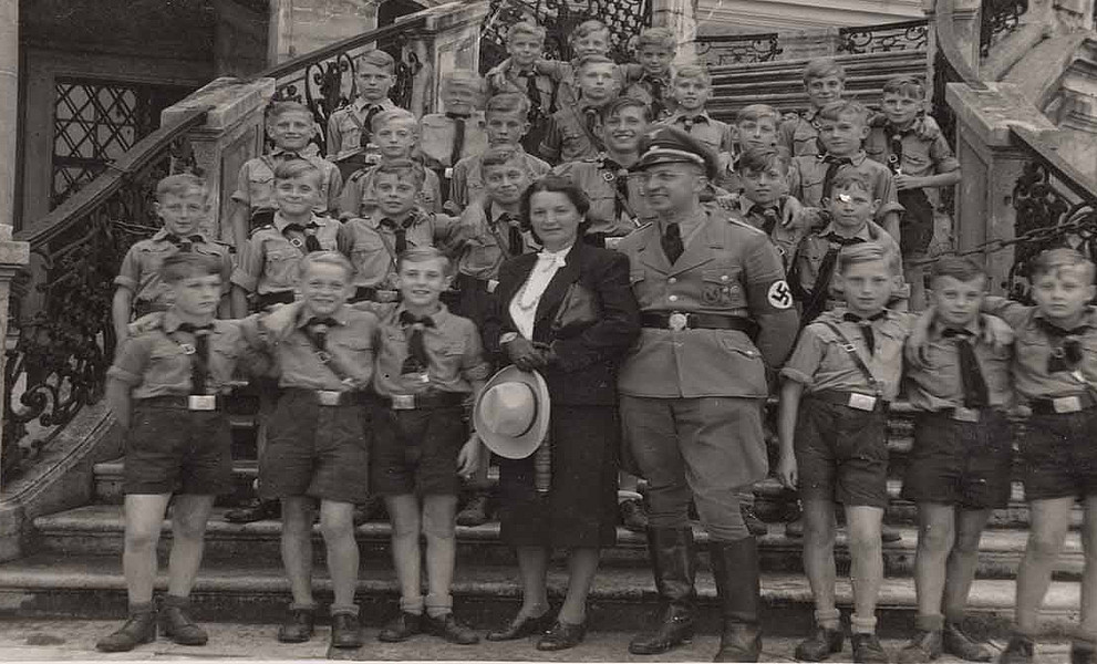 Gruppe von Jungen in Uniform, eine Frau und ein Mann in NS-Uniform stehen auf einer Treppe vor einem Gebäude, alle blicken in die Kamera.