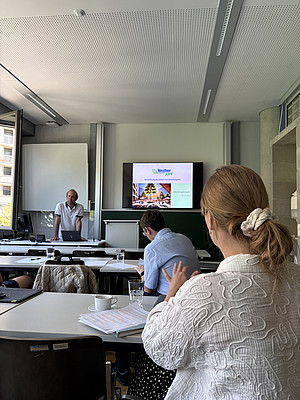 Blick in einen Seminarraum während einer Präsentation. Im Vordergrund sitzt eine Teilnehmerin, weitere Personen sitzen an Tischen. Vorne steht ein Vortragender neben einem Bildschirm, auf dem eine Präsentation zum Projekt „Weatherpark“ zu sehen ist. ©Paul Schweighofer