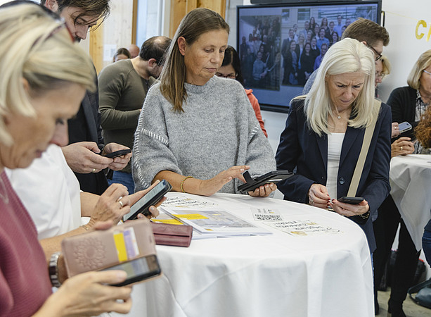 Mehrere Gäste nutzen Smartphones an einem Tisch mit Unterlagen und einem QR-Code; im Hintergrund ist ein Gruppenfoto auf einem Bildschirm zu sehen. ©@foto-gasser.at