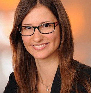 Frau Wohlmuth trägt eine Brille und steht vor einem orangen Hintergrund