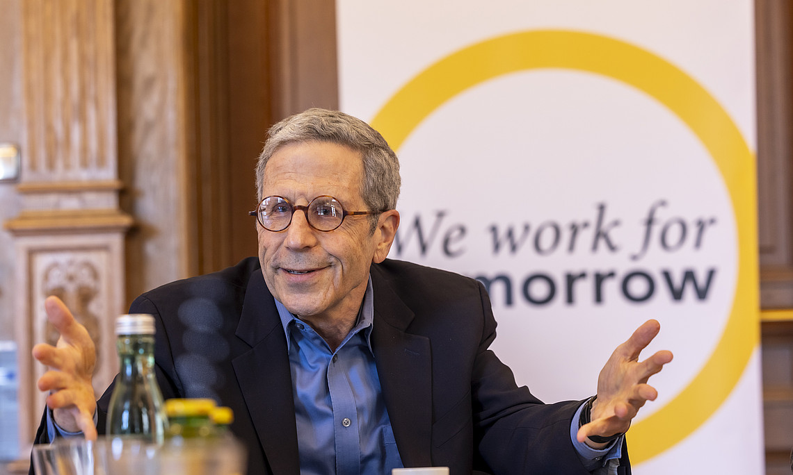 A photograph of an elegant man in his seventies, wearing glasses and with short hair, sitting at a table and talking to others. Behind him is a wall on which the text "We work for tomorrow" is written.