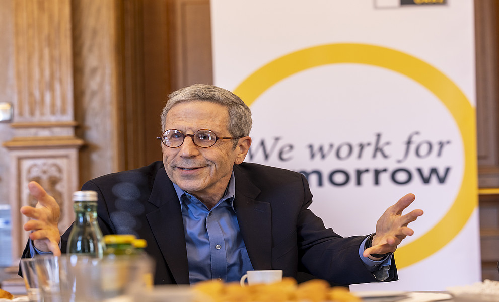Ein Foto von einem eleganten Mann in den Siebzigern, der eine Brille trägt und kurze Haare hat, an einem Tisch sitzt und mit anderen spricht. Hinter ihm befindet sich eine Wand, auf der der Text "We work for tomorrow" steht.