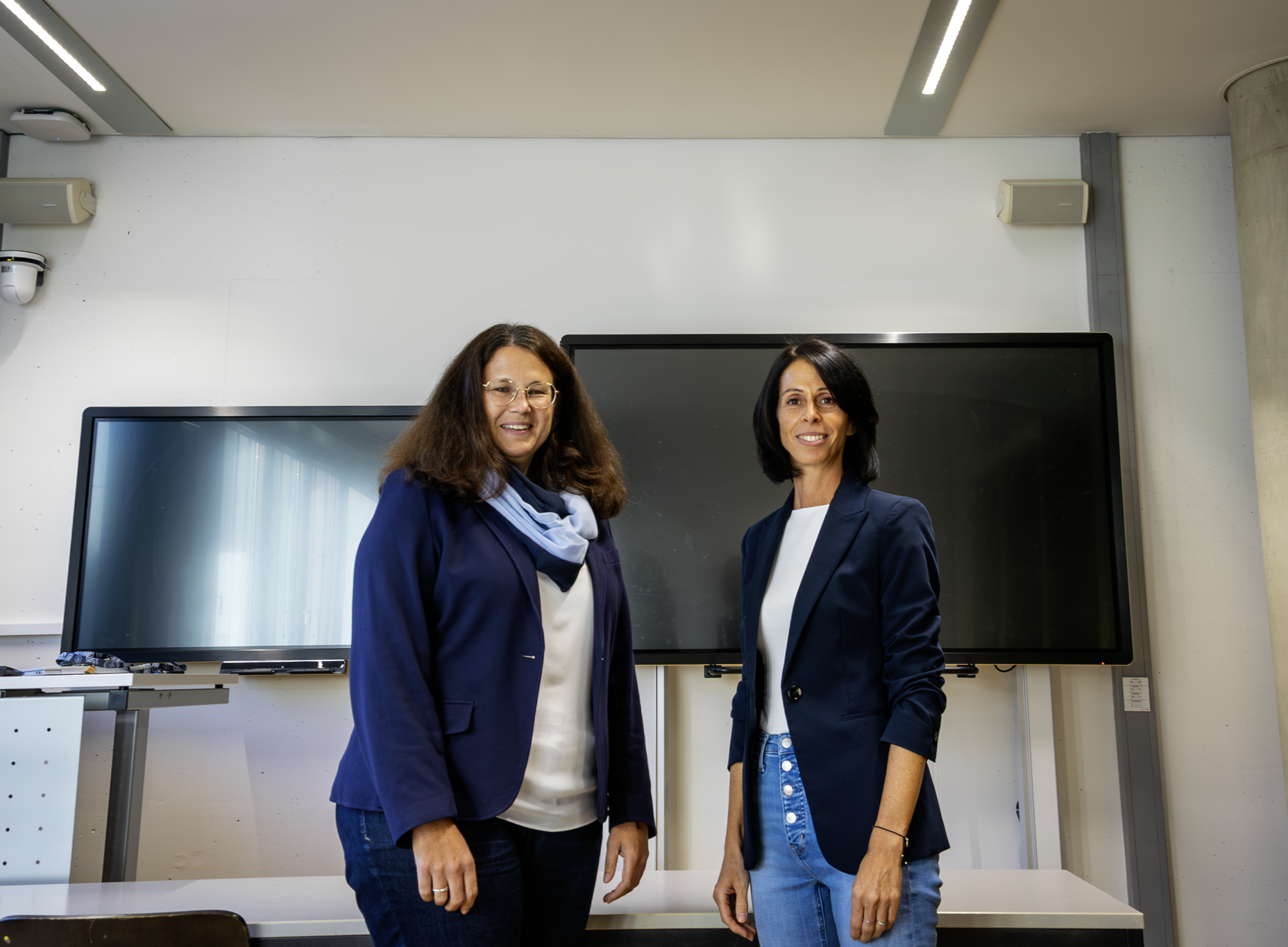Zwei Personen stehen lächelnd in einem modernen Seminarraum vor zwei großen Bildschirmen. Beide tragen dunkle Blazer und helle Oberteile; im Hintergrund sind Möbel und technische Ausstattung des Unterrichtsraums zu sehen. ©SOWI/Polutak
