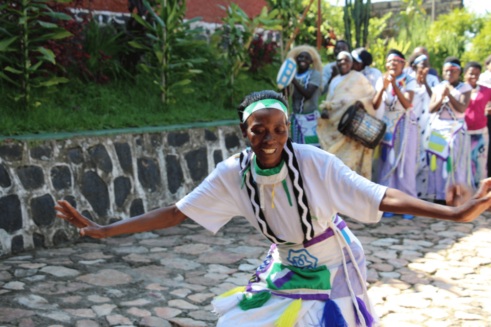 Fotografie einer tanzenden und lachenden ruandischen Frau in traditionellem Gewand bei einer Festlichkeit; im Hintergrund sind weitere Personen zu sehen, die mitklatschen und teilweise auf Trommeln spielen.
