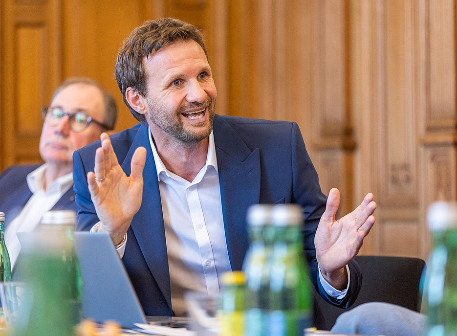 Das Foto zeigt einen Geschäftsmann in blauem Anzug und weißem Hemd, der mit einem Laptop an einem Tisch sitzt und sich in einem Konferenzraum mit anderen Personen unterhält, während er lächelt und gestikuliert. Auf dem Schreibtisch stehen grüne Glasflaschen, und die Wände sind mit Holzpaneelen verkleidet. Die Beleuchtung ist natürlich. ©www.photoworkers.at