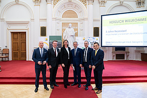 A group of people in formal attire stand on a red carpet in a magnificent hall with high, ornate walls and a statue in the background. On the right-hand side of the picture, a large screen can be seen with the words “Welcome to the Graz Economic Talks” and further information. There are also banners and a wooden door in the background.