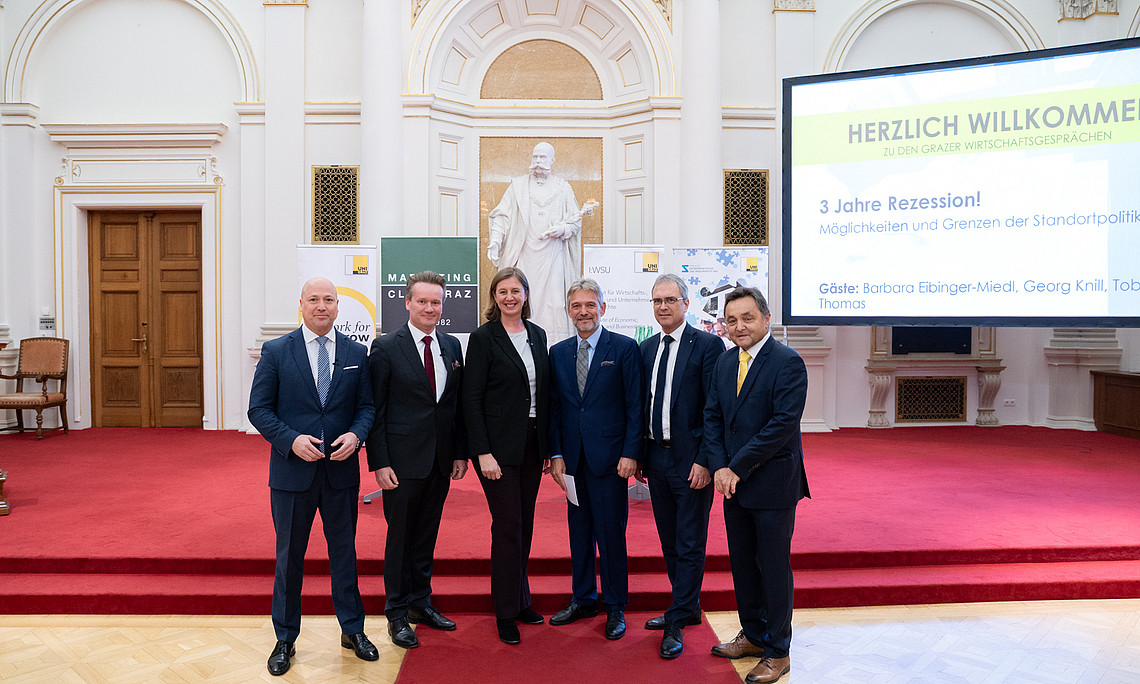 A group of people in formal attire stand on a red carpet in a magnificent hall with high, ornate walls and a statue in the background. On the right-hand side of the picture, a large screen can be seen with the words “Welcome to the Graz Economic Talks” and further information. There are also banners and a wooden door in the background.