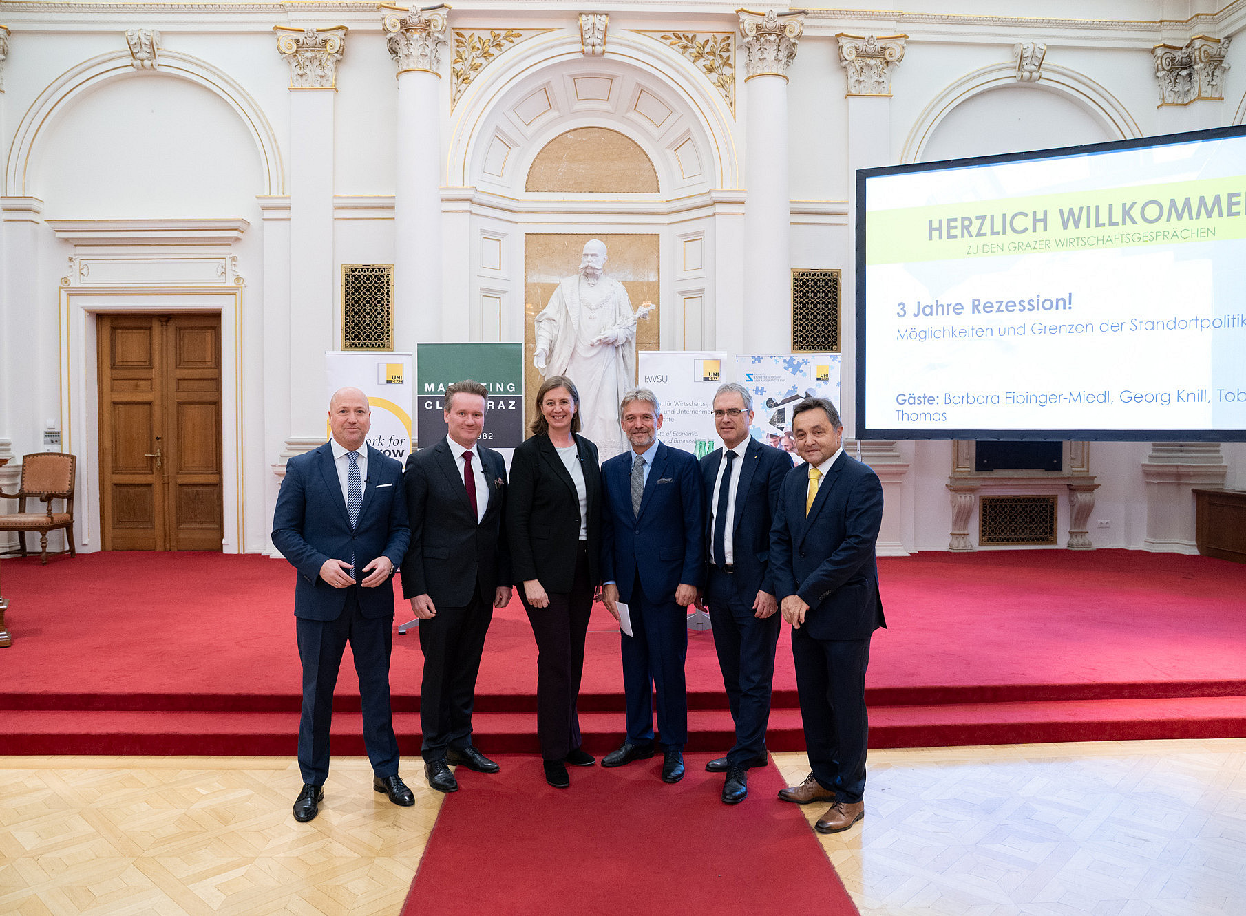 Eine Gruppe von Personen in formeller Kleidung steht auf einem roten Teppich in einem prunkvollen Saal mit hohen, verzierten Wänden und einer Statue im Hintergrund. Im rechten Bereich des Bildes ist eine große Leinwand mit der Aufschrift „Herzlich Willkommen zu den Grazer Wirtschaftsgesprächen“ und weiteren Informationen sichtbar. Im Hintergrund befinden sich zudem Banner und eine Holztür. 