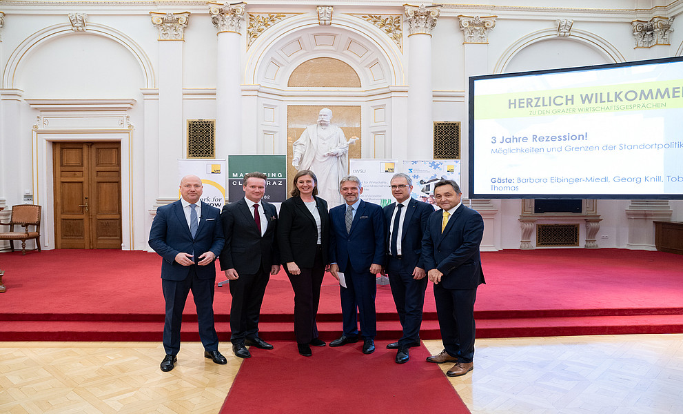 Eine Gruppe von Personen in formeller Kleidung steht auf einem roten Teppich in einem prunkvollen Saal mit hohen, verzierten Wänden und einer Statue im Hintergrund. Im rechten Bereich des Bildes ist eine große Leinwand mit der Aufschrift „Herzlich Willkommen zu den Grazer Wirtschaftsgesprächen“ und weiteren Informationen sichtbar. Im Hintergrund befinden sich zudem Banner und eine Holztür.