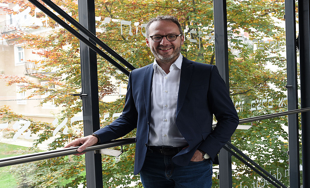 Ralf Lutz vor einer Glaswand, dahinter ein Baum mit herbstlichem Laub