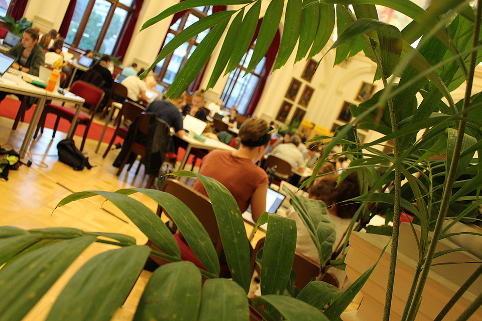 Menschen beim Schreiben in der Aula im Hintergrund. Im Vordergrund sind Palmenblätter.