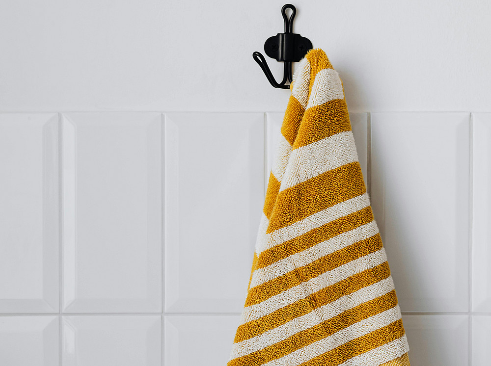Ein weiß-gelb gestreiftes Handtuch hängt an einem schwarzen Haken an einer weiß gefliesten Wand. ©pexels - Karolina Grabowska