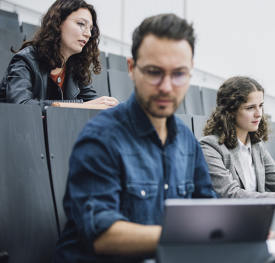 Ein Studierender schaut im Hörsaal konzentriert auf seinen Laptop, während hinter ihm zwei Studierende mit Notizbuch dem Vortrag zuhören.