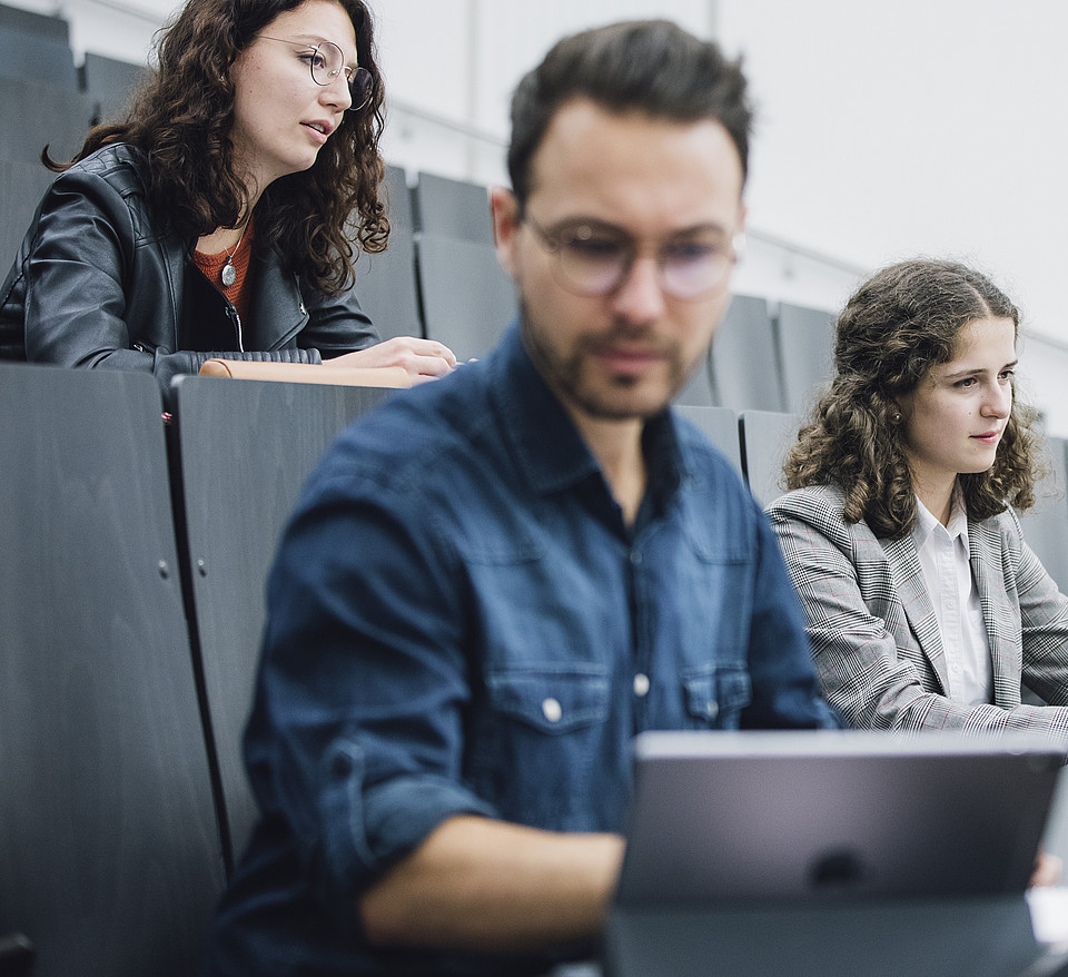 Ein Studierender schaut im Hörsaal konzentriert auf seinen Laptop, während hinter ihm zwei Studierende mit Notizbuch dem Vortrag zuhören.