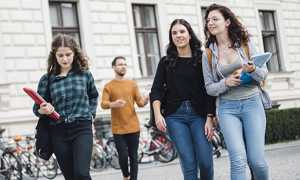 Studierende gehen im Freien vor den Fahrradständern vor dem Hauptgebäude der Uni Graz und unterhalten sich miteinander.