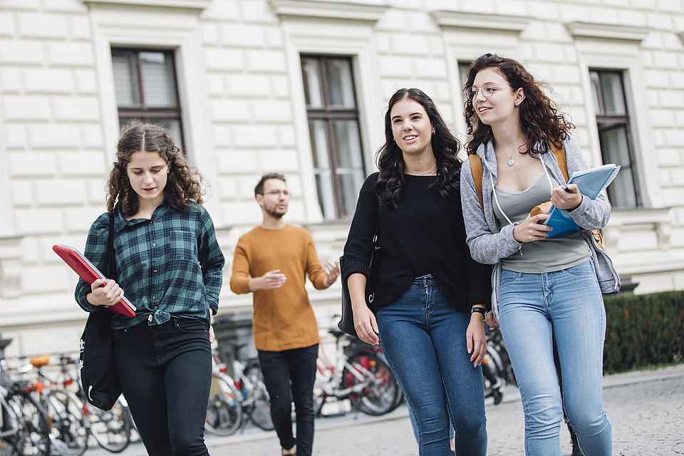 Studierende gehen im Freien vor den Fahrradständern vor dem Hauptgebäude der Uni Graz und unterhalten sich miteinander.