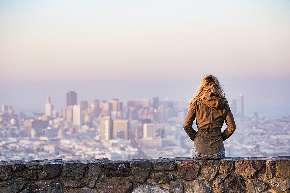 Eine Studierende sitzt auf einer Mauer und blickt auf eine Stadt