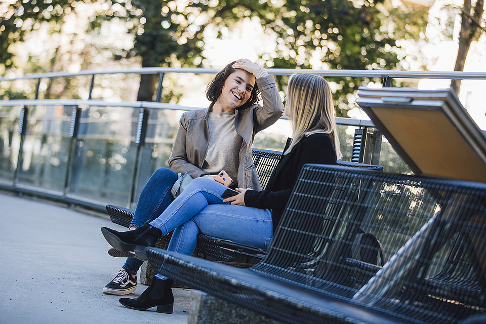 Zwei Studierende sitzen am Campus auf einer Bank und unterhalten sich