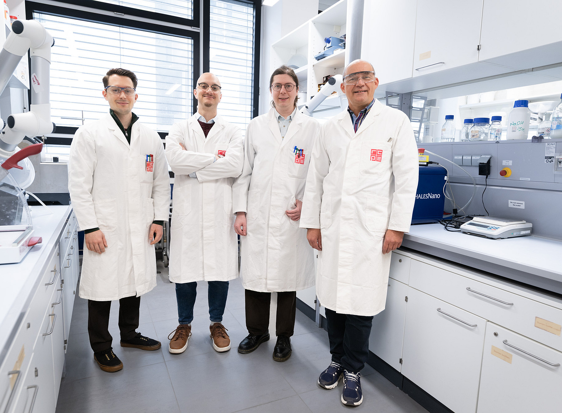 Four people in white lab coats are standing side by side in a bright chemistry laboratory. Their faces have been obscured. They are looking towards the camera; three have their arms hanging by their sides, whilst one person has their arms crossed. To the left and right, laboratory equipment, shelves, cables and several labelled bottles can be seen; daylight streams into the laboratory through large windows in the background. ©Uni Graz/Angele