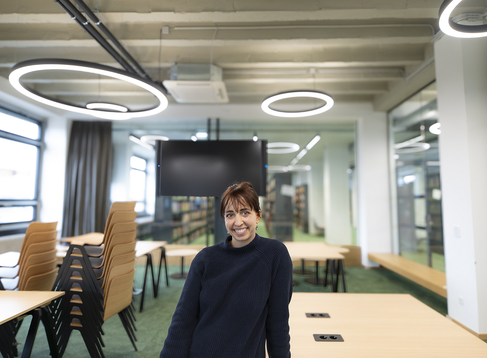 Modern seminar or study room with large windows, stacked wooden chairs on tables and bright ring lamps on the ceiling; in the background is a large screen, behind which more bookshelves are visible. ©Uni Graz/Tzivanopoulos
