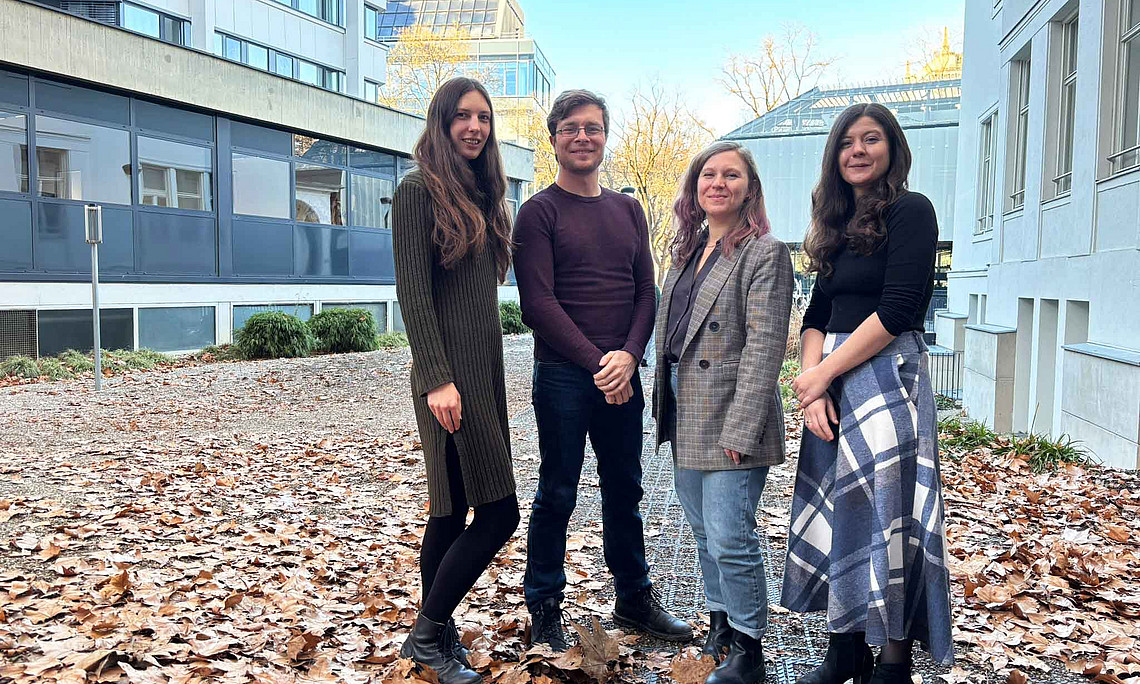 Vier junge Erwachsene stehen auf herbstbedecktem Boden zwischen modernen Gebäuden; sie tragen Jacken, Pullover, Kleid, Rock und Stiefel.