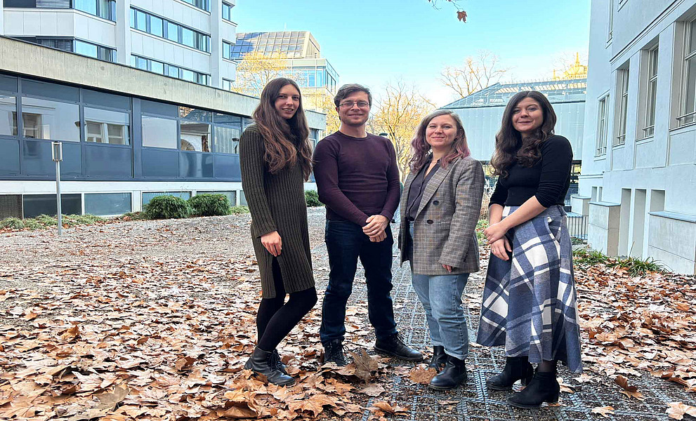 Vier junge Erwachsene stehen auf herbstbedecktem Boden zwischen modernen Gebäuden; sie tragen Jacken, Pullover, Kleid, Rock und Stiefel.