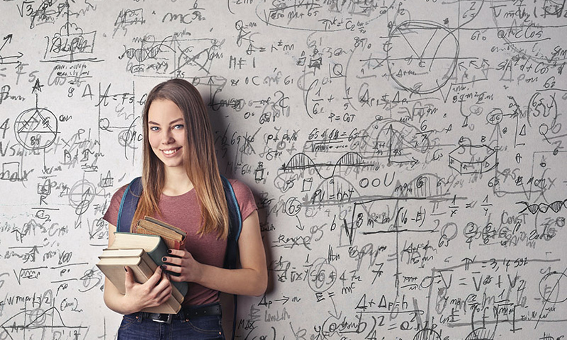 Lächelndes Mädchen mit Schulbüchern in der Hand steht vor einer Tafel mit mathematischen Formeln