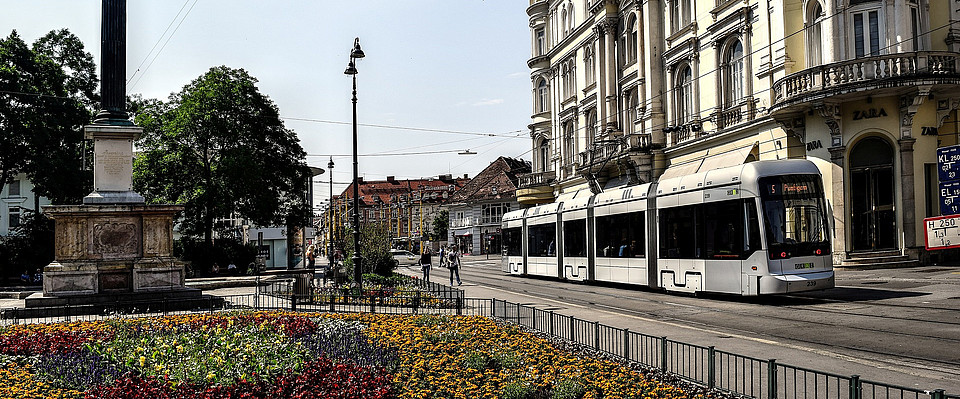 Aufnahme der Grazer Innenstadt mit einer Straßenbahn, historischen Gebäuden und einer Statue mit Brunnen